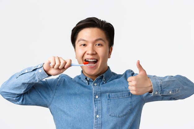 Orthodontics, dental care and hygiene concept. Close-up of satisfied happy asian man brushing teeth with braces, holding toothbrush and showing thumbs-up in approval, white background