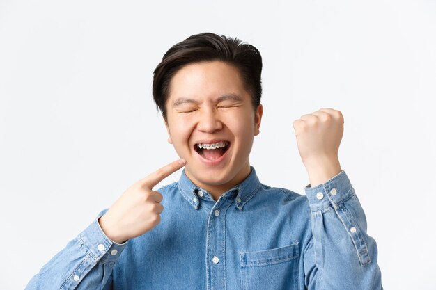 歯列矯正と口腔病学の概念。新しい歯ブレースを喜んで、口を指して、笑顔、拳ポンプ、白い背景に勝利する興奮して幸せなアジア人の男のクローズアップ
