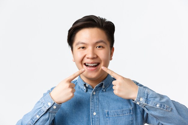 Orthodontics and stomatology concept. Close-up of happy smiling asian man pointing fingers at dental braces on teeth with pleased expression, recommend dentist clinic, standing white wall