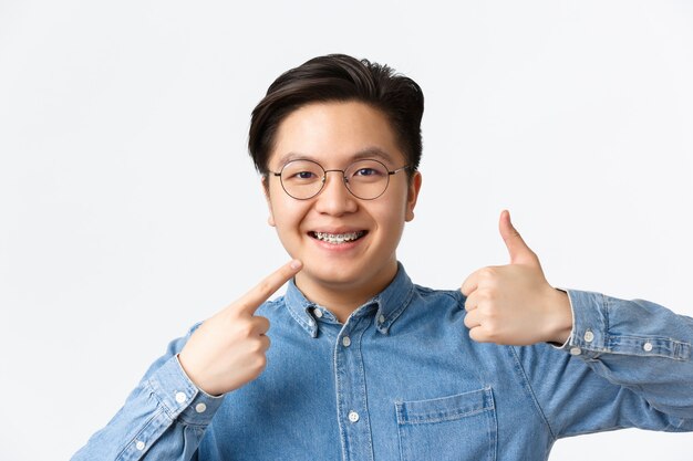 Orthodontics and stomatology concept. Close-up of satisfied asian guy, dental clinic client smiling happy and pointing at his dental braces and showing thumbs-up in approval, recommend