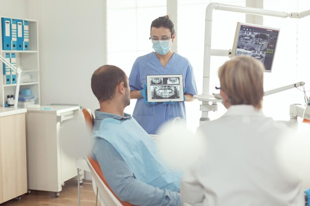치아 방사선 사진이 있는 디지털 태블릿을 들고 있는 치열 교정 간호사는 치통을 예방하기 위해 아픈 사람의 구강 치료에 대해 설명합니다. 현대 치과 사무실에서 치과 의자에 앉아있는 환자