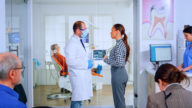 Free photo orthodontist using tablet explaining dental x ray to patient standing in waiting area of stomatologic office. dentist showing to woman radiography using digital device working in modern clinic.