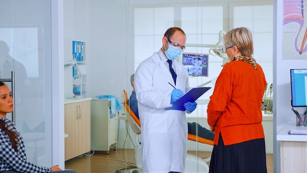 Orthodontist with mask speaking with elderly woman standing in waiting area of stomatological clinic taking notes on clipboard. Nurse typing on computer appointments in modern crowded office