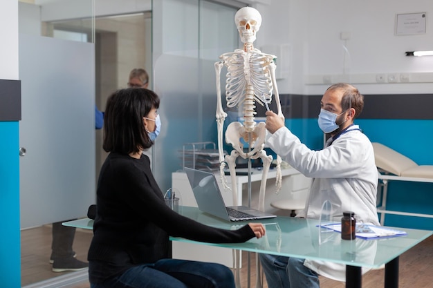 의료 캐비닛에 있는 여성에게 인간 골격의 뼈 부상을 설명하는 정형외과 전문의. 해부학적 구조와 진단을 보여주기 위해 정골의학 모델에서 척수를 가리키는 의사.
