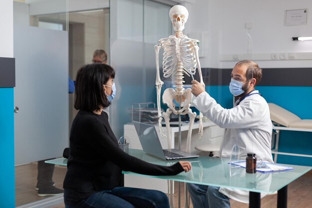Orthopedic specialist explaining bones injury on human skeleton to woman in medical cabinet. Physician pointing at spinal cord on osteopathy model to show anatomy structure and diagnosis.