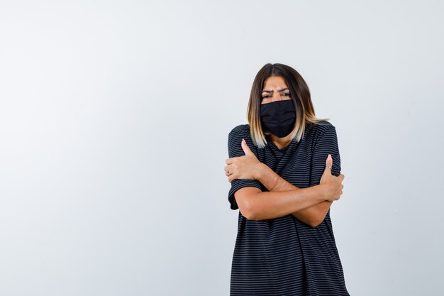 Ortrait of lady hugging herself or feeling cold in black dress, medical mask and looking upset front view