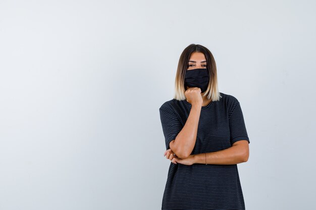 검은 드레스, 의료 마스크에 주먹에 턱을지지하고 현명한 정면보기를 보는 숙녀의 Ortrait