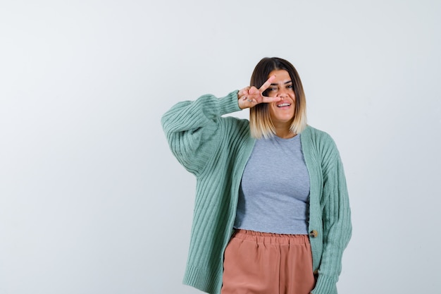 Ortrait of woman showing V-sign on eye in casual clothes and looking joyful front view