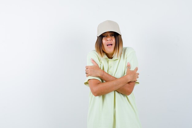 Ortrait of young female hugging herself in t-shirt, cap and looking chilled front view