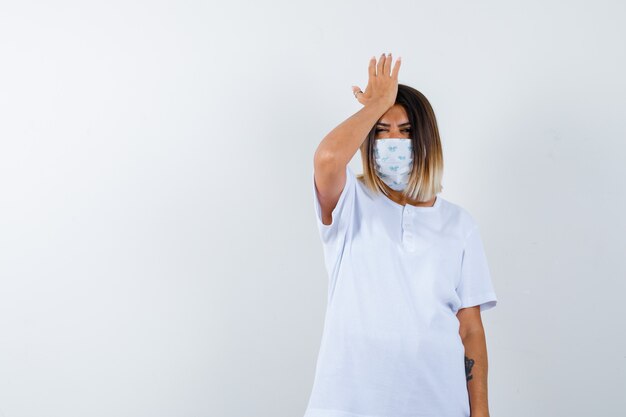 Ortrait of young female keeping hand on head in t-shirt, mask and looking forgetful front view