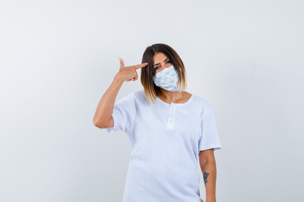 Ortrait of young female pointing at head in t-shirt, mask and looking thoughtful front view