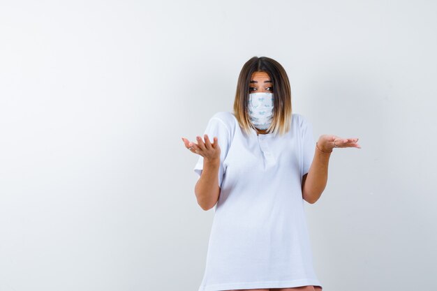 Ortrait of young female showing helpless gesture in t-shirt, mask and looking puzzled front view