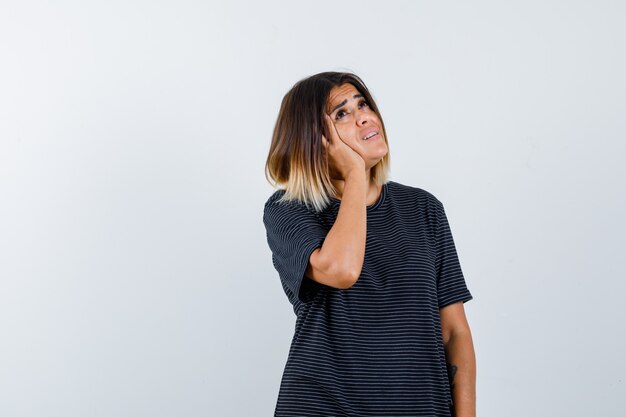 Ortrait of young lady holding hand on cheek while looking up in polo dress and looking confused front view