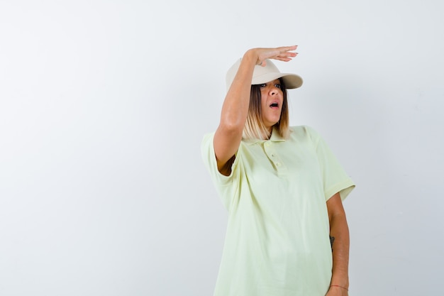 Free photo ortrait of young lady holding hand on head to see clearly in t-shirt, cap and looking puzzled front view