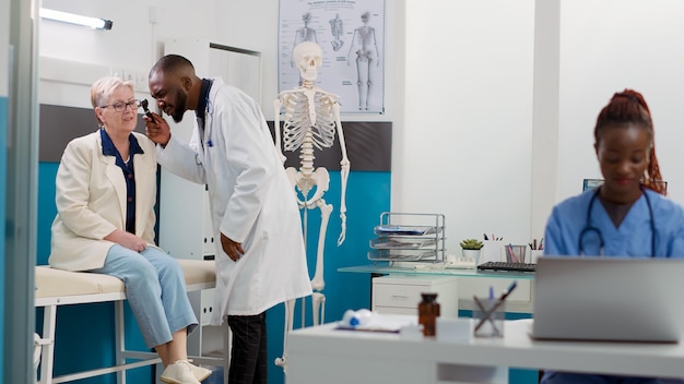 Otology specialist using otoscope to do ear examination with senior woman in cabinet, consulting patient with otolaryngology instrument. Doing ent consultation for audiology diagnosis.