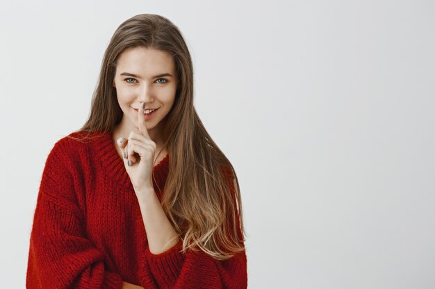 Our dirty little secret. Studio shot of pleased good-looking caucasian girlfriend in red loose sweater, saying shh with shush gesture, holding index finger over mouth and smiling flirty over gray wall