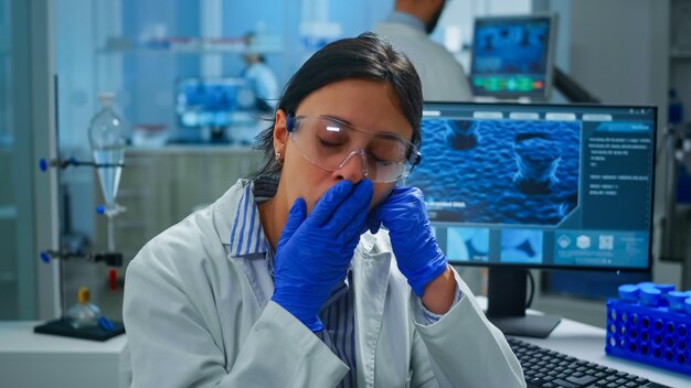 발발 화학자는 카메라에 피곤 찾고 하품을 갖춘 현대적인 실험실에 앉아 있습니다. 과학 연구, 백신 개발을 위한 첨단 화학 도구를 사용하여 바이러스 진화를 조사하는 과학자