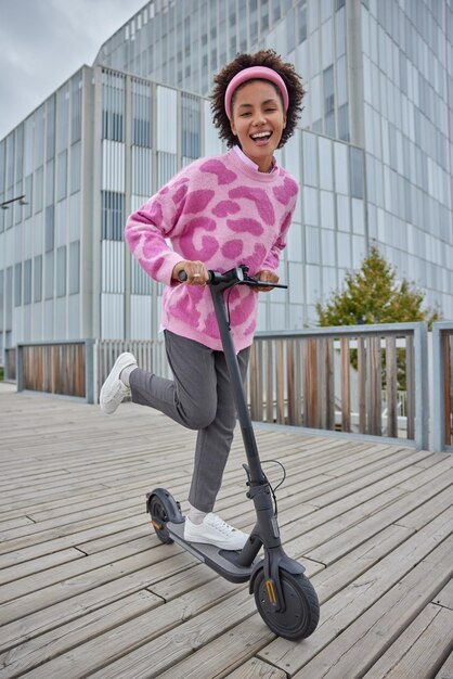 Outdoor activities lifestyle and ecological transport concept. Joyful curly haired young female model rides electric scooter has fun enjoys awesome ride during leisure time poses in urban place