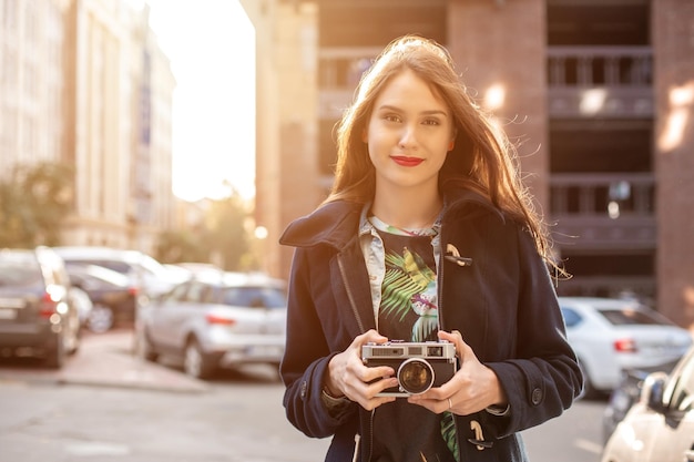 Outdoor autumn smiling lifestyle portrait of pretty young woman, having fun in the city with camera, travel photo of photographer. Making pictures in hipster style. Sun flare