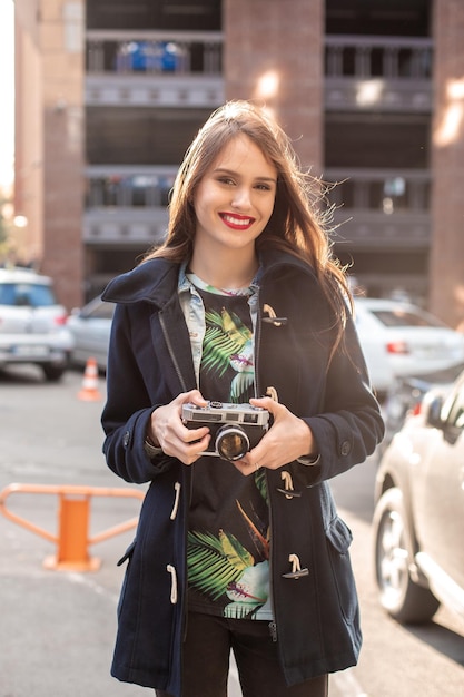 Outdoor autumn smiling lifestyle portrait of pretty young woman, having fun in the city with camera, travel photo of photographer. Making pictures in hipster style.