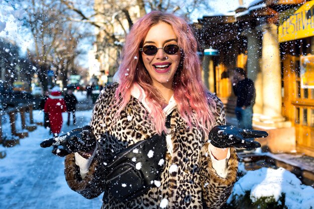 특이한 분홍색 머리카락을 가진 예쁜 여자의 야외 쾌활한 라이프 스타일 초상화, 트렌디 한 바디 표범 모피 재킷, 빈티지 90 년대 스타일의 선글라스와 부랑자 가방, 그런지 거리 착용, 눈이 내리는 도시.