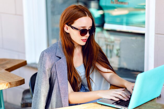 Outdoor city fashion portrait of young businesswoman working at cafe on terrace at sunny day, casual stylish outfit, mint details, using her laptop, cafe break, business concept.