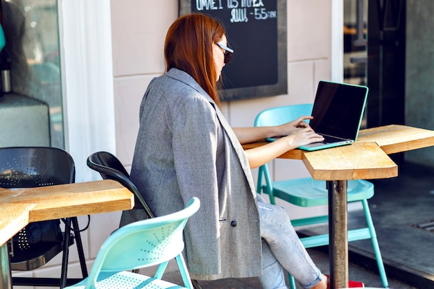 Ritratto di moda città all'aperto di giovane donna di affari che lavora al caffè sulla terrazza al giorno pieno di sole, vestito elegante casual, dettagli di menta, utilizzando il suo computer portatile, pausa caffè, concetto di affari.