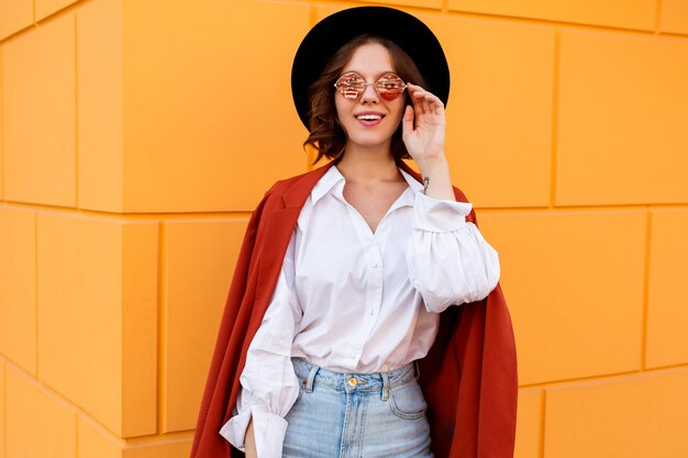 Outdoor close up portrait of blissful brunette short haired female posing over yellow wall. Trendy hat, pink glasses, white blouse and jeans.