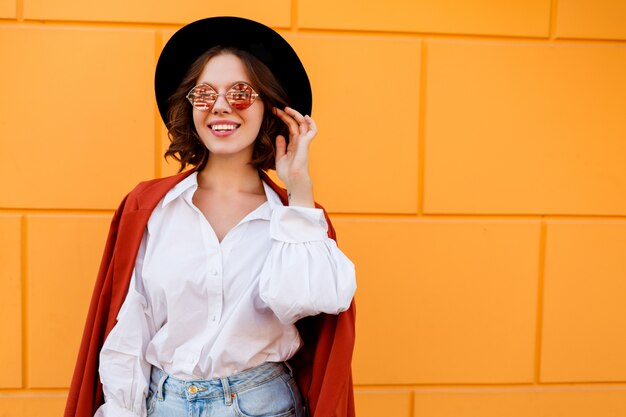 Outdoor close up portrait of blissful brunette short haired female posing over yellow wall. Trendy hat, pink glasses, white blouse and jeans.