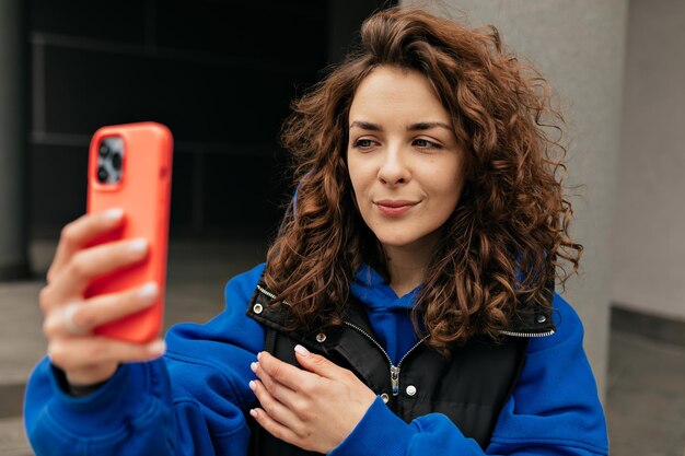 青いセーターと暗いジャケットの巻き毛の素敵な女性の屋外クローズアップの肖像画は、スマートフォンで自分撮りを作っています