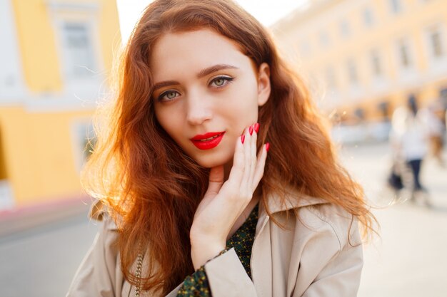 Outdoor close up portrait of lovely ginger woman with wavy hairs in beige coat. Red lips and nail.