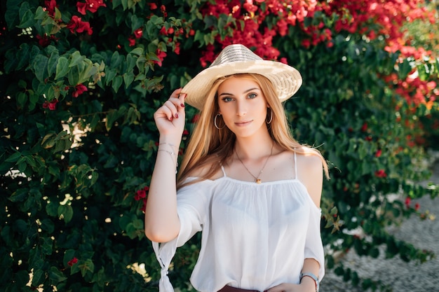 Outdoor close up portrait of young beautiful happy smiling curly girl wearing stylish straw hat in street near blooming roses. Summer fashion concept. Copy space