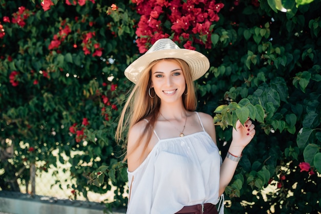 Outdoor close up portrait of young beautiful happy smiling curly girl wearing stylish straw hat in street near blooming roses. Summer fashion concept. Copy space