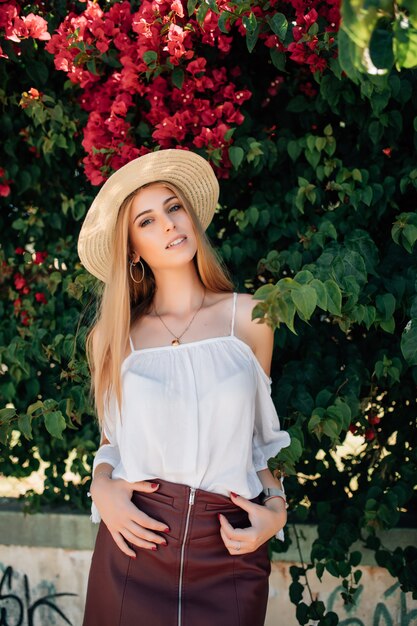 Outdoor close up portrait of young beautiful happy smiling curly girl wearing stylish straw hat in street near blooming roses. Summer fashion concept. Copy space