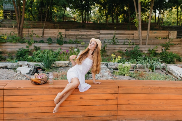 Free photo outdoor of dreamy barefooted lady with long curly hair sitting on wooden bench in park and looking away. romantic girl in straw hat and white dress posing in front of flowerbed.