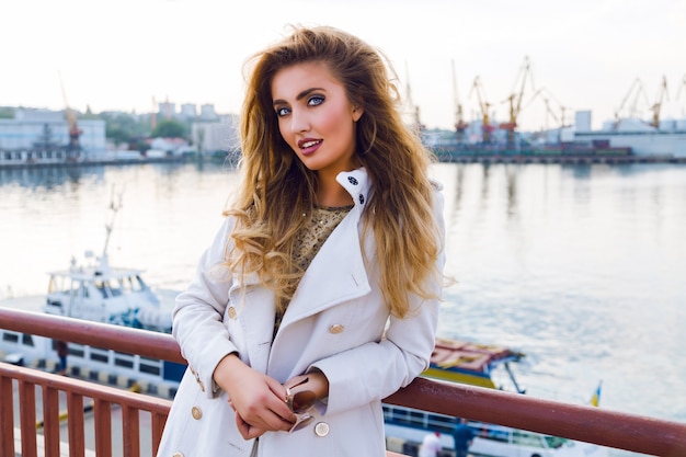 Outdoor fall fashion portrait of sexy elegant lady posing neat sea port dreaming and thinking, wearing white cashmere white coat have curled hairs and bright makeup. Evening sunlight, soft colors.