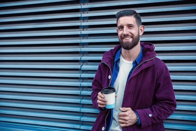 Free photo outdoor fall or winter portrait of handsome hipster man with beard, white t-shirt, blue shirt and maroon jacket holding cup of hot coffee. ribbed urban wall background.