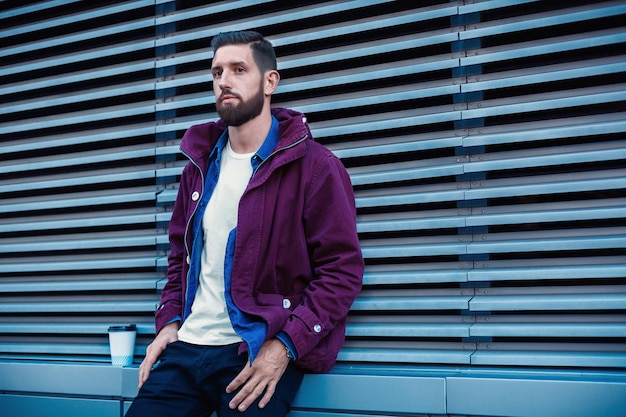 Free photo outdoor fall or winter portrait of handsome hipster man with beard, white t-shirt, blue shirt and maroon jacket holding cup of hot coffee. ribbed urban wall background.