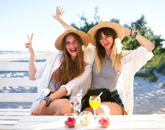 2人の姉妹の屋外のファニーの肖像画は、ビーチバー、自由奔放に生きる流行に敏感な服、おいしいカクテルを飲んで、夏の海の休暇で笑顔としかめっ面をして楽しんでいる友人の女の子を打つ。