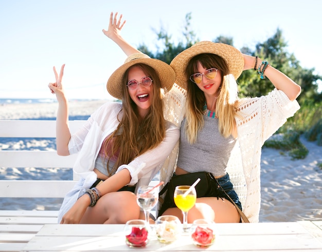 Outdoor  fanny portrait of two sister beat  friends girl having fun hugs smiling and making grimaces on beach bar, boho hipster clothes, drinking tasty cocktails, summer ocean vacation .