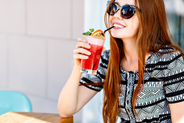 Foto gratuita ritratto di bellezza alla moda all'aperto di signora elegante glamour, capelli lunghi incredibili, abito vintage di lusso e occhiali da sole a occhi di gatto, bere gustosi cocktail freddi, terrazza del caffè della città, viaggi, gioia, relax.