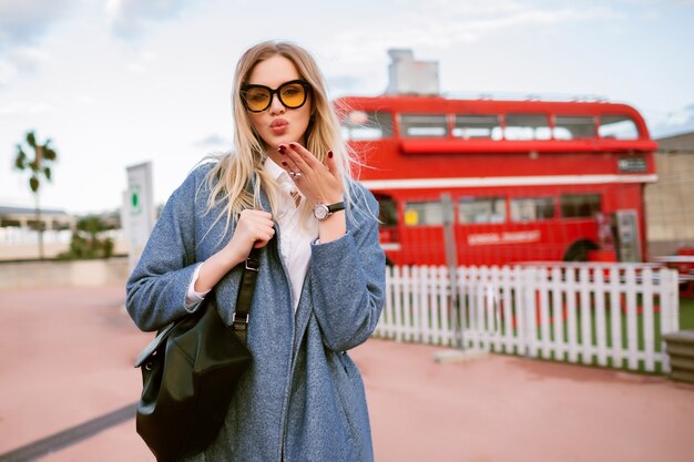 Outdoor fashion image of stylish young woman posing at London street , elegant casual business outfit, sending kiss and looking on camera , autumn spring mid season time, toned colors.