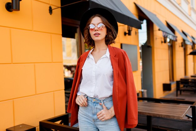 Outdoor fashion image of young elegant woman in orange jacket and white blouse walking in sunny city during weekends.