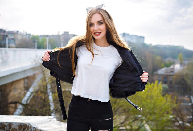 Outdoor fashion portrait of cute blonde woman smiling and having fun, long hairs, bright make up, biker leather jacket, street fashion.