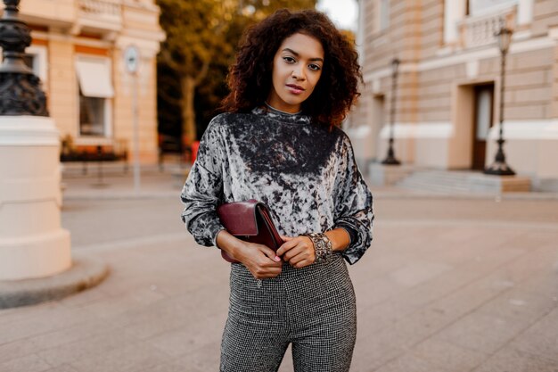 トレンディな秋の服、灰色のベルベットのセーター、高級財布を着て魅力的な官能的な若いスタイリッシュな黒人女性のアウトドアファッションの肖像画。