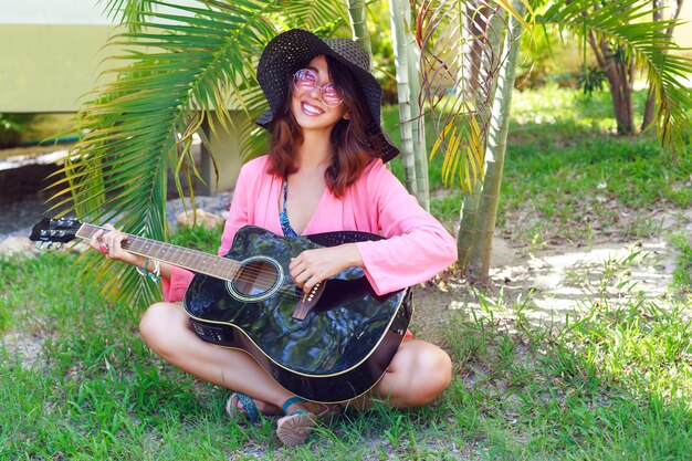 Outdoor fashion portrait of happy pretty smiling hippie woman sitting at the grass and holding acoustic guitar. Hot tropical country, green background. Summer outfit with hat and pink sunglasses.