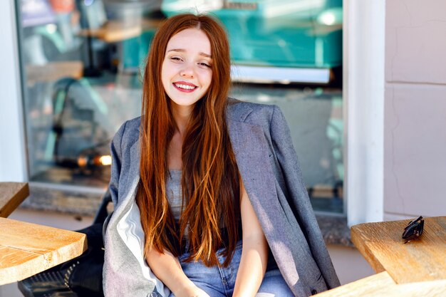 Outdoor fashion portrait of stylish ginger woman, posing at terrace cafe, at sunny day, wearing boyfriend jacket, bright fresh colors.