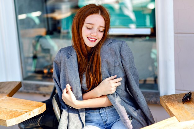 Outdoor fashion portrait of stylish ginger woman, posing at terrace cafe, at sunny day, wearing boyfriend jacket, bright fresh colors.