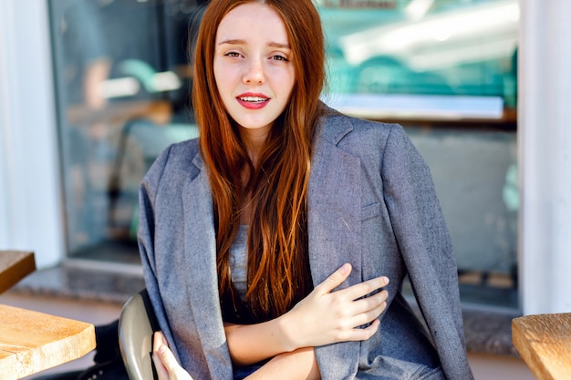 Outdoor fashion portrait of stylish ginger woman, posing at terrace cafe, at sunny day, wearing boyfriend jacket, bright fresh colors.