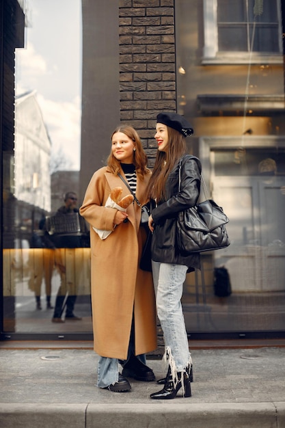 Outdoor fashion portrait of two young beautiful women wearing trendy stylish clothes. Young girls posing in street of european city. Girl holding a baguettes in kraft paper bag.